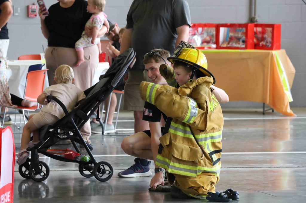A young child dressed in a firefighter costume smiles while kneeling beside an older boy, who is crouching down and engaging with him. In the background, families are gathered, with some children playing nearby.