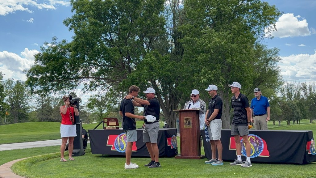 A group of golfers receives an award during a ceremony at a golf course, with a podium in the foreground and a camera operator capturing the event.