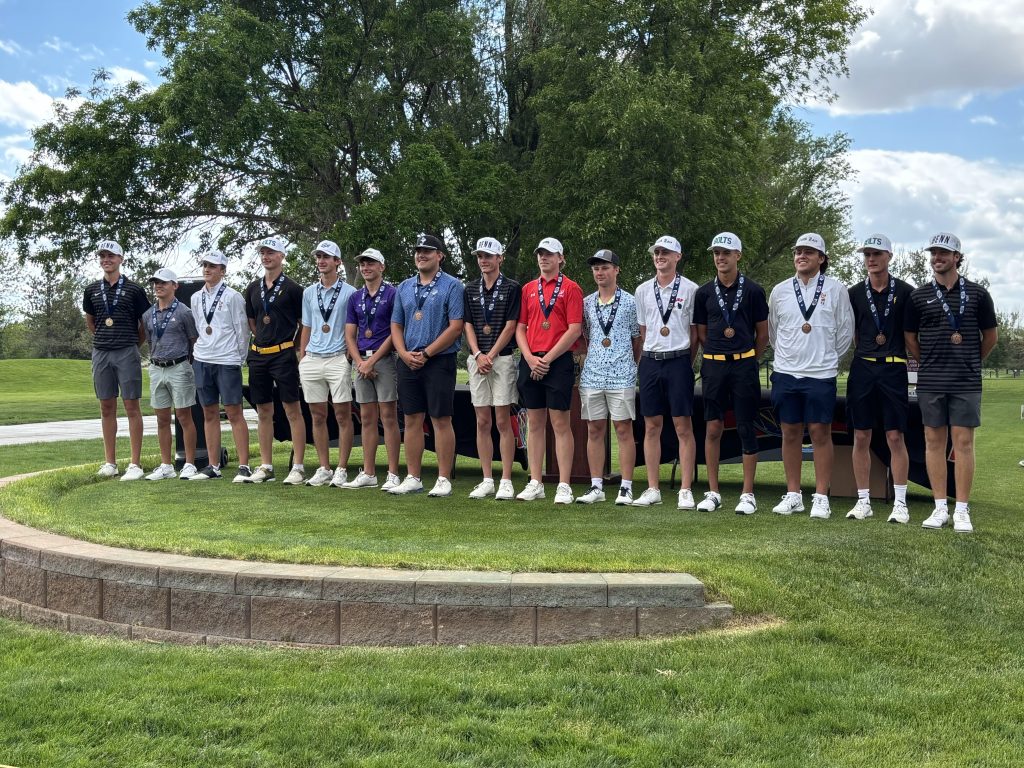 Group of 15 male golfers standing in a line, wearing golf attire and medals, on a grassy area during a tournament ceremony.