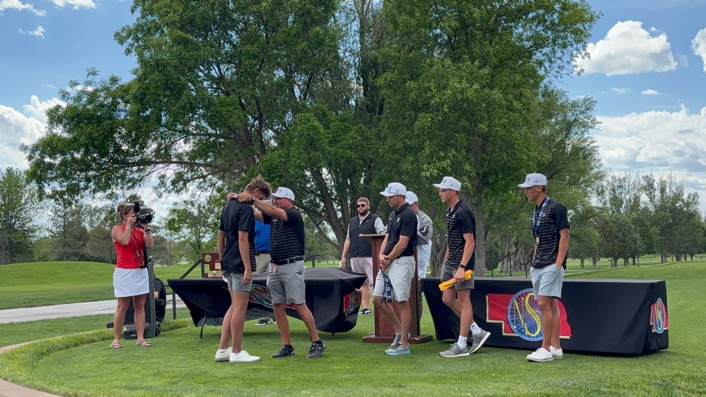 Group of golfers at an award ceremony on a sunny day, with a woman filming, a trophy on a table, and a backdrop showcasing the event logo.