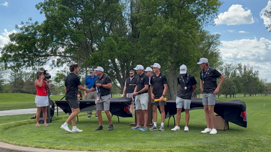 A group of men in golf attire stand together on a golf course, receiving awards. One man is shaking hands with an official while a woman in a red shirt and another man film the event.