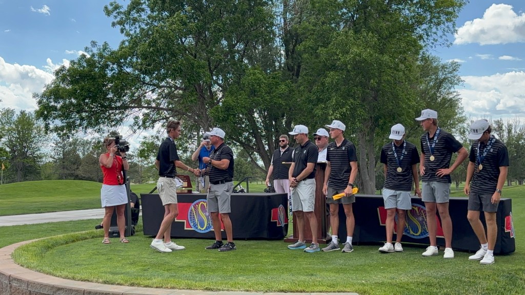 A group of golfers on stage receiving awards at a tournament ceremony, with one golfer shaking hands with an official and a camera person nearby.