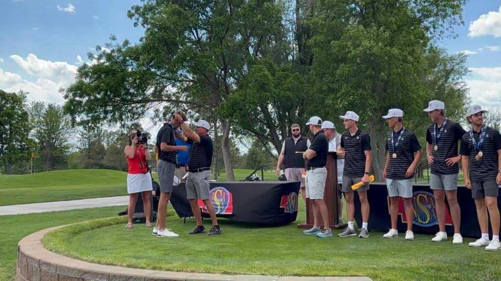 A group of male golfers wearing matching shirts and medals stands on a grassy podium during an awards ceremony. One golfer is being embraced and congratulated by another, while a female photographer captures the moment.