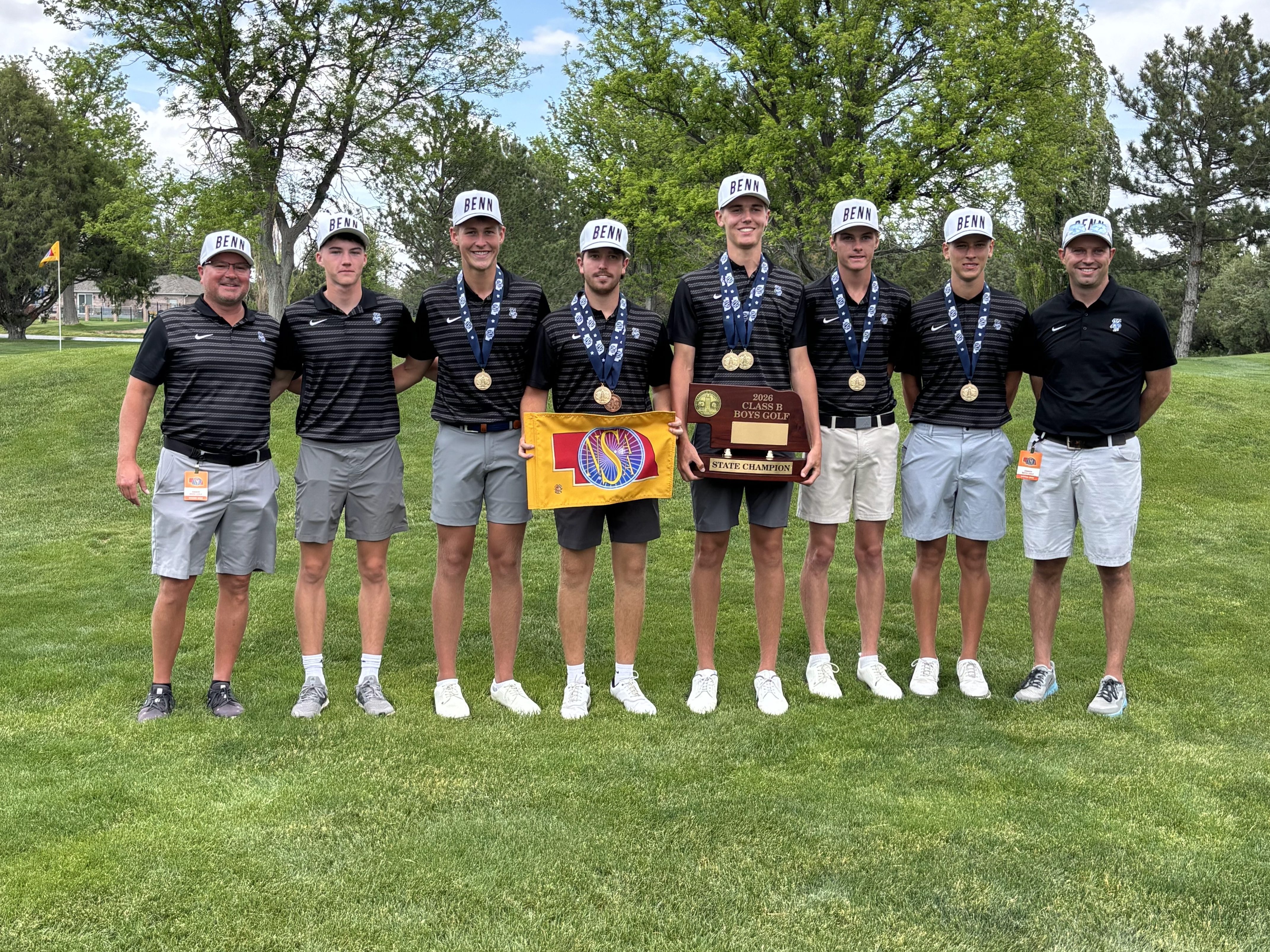 A group of seven individuals stands on a golf course, celebrating a championship win. They are wearing matching black and white striped shirts and hats, holding medals and a trophy. The backdrop features green grass and trees under a partly cloudy sky.
