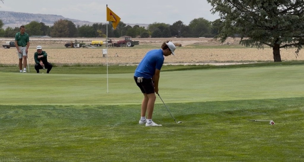 A golfer in blue prepares to putt on the green while two spectators watch in the background. A flag is visible next to the hole, with a rural landscape beyond.