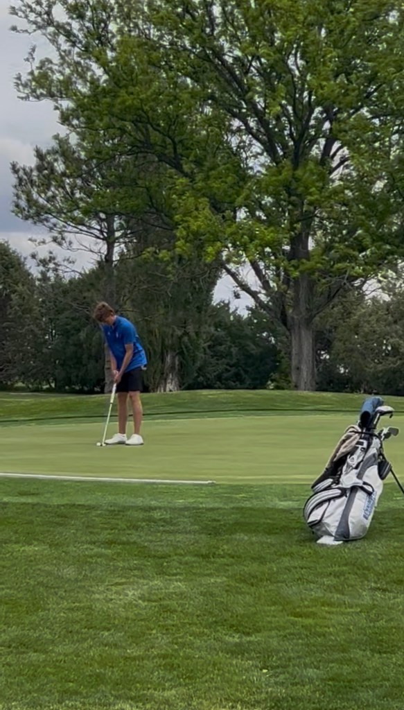 A young golfer in a blue shirt and black shorts preparing to putt on a green golf course, with a golf bag in the foreground.