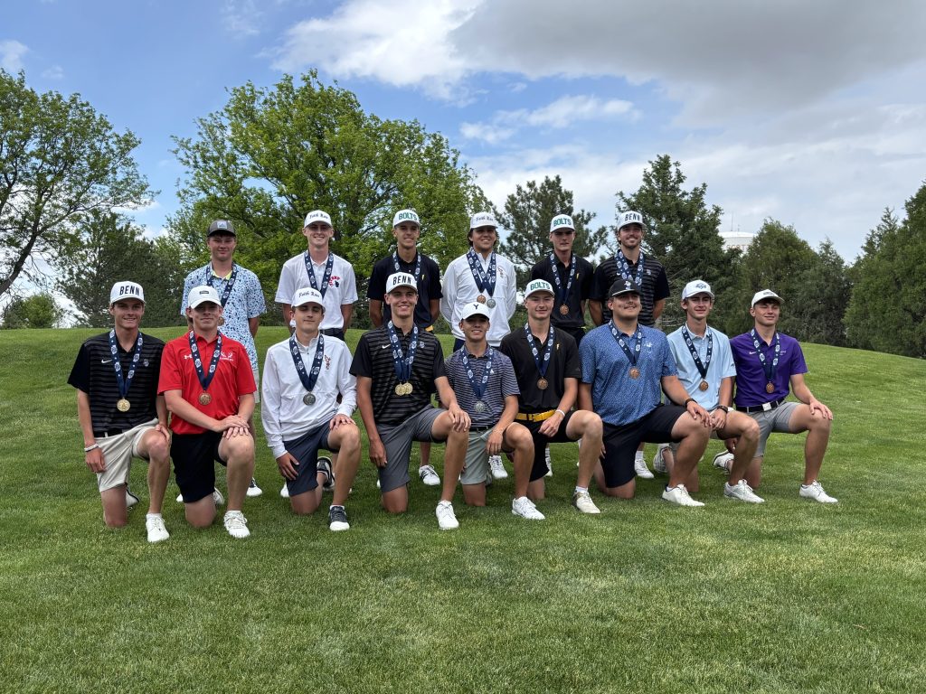 Group photo of 16 young male golfers wearing various colored shirts and hats, all smiling while wearing medals around their necks, posing on a grassy area with trees in the background.