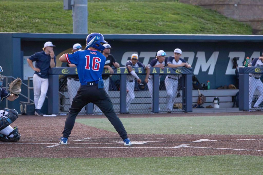 A baseball player wearing a blue jersey with the number 16 stands at the plate, preparing to bat. A catcher is positioned behind home plate, while players from the dugout look on from the background.
