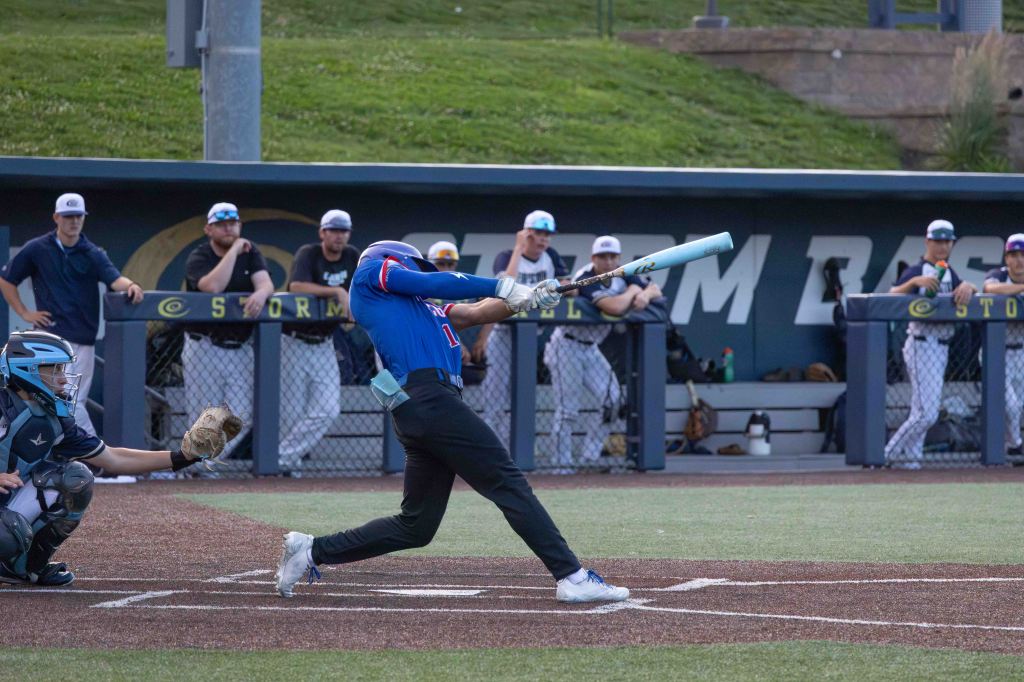 A baseball player swings a bat during a game, with a catcher poised to receive a pitch. Teammates and coaches watch from the dugout in the background.
