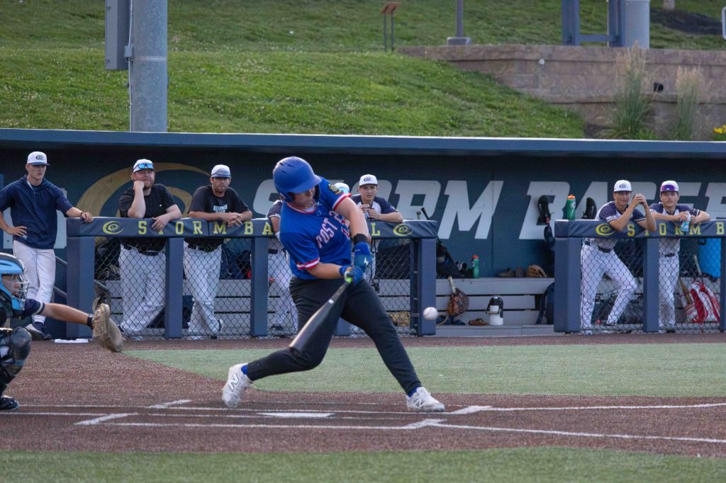 A baseball player swings at a pitch during a game, with teammates observing from the dugout in the background.