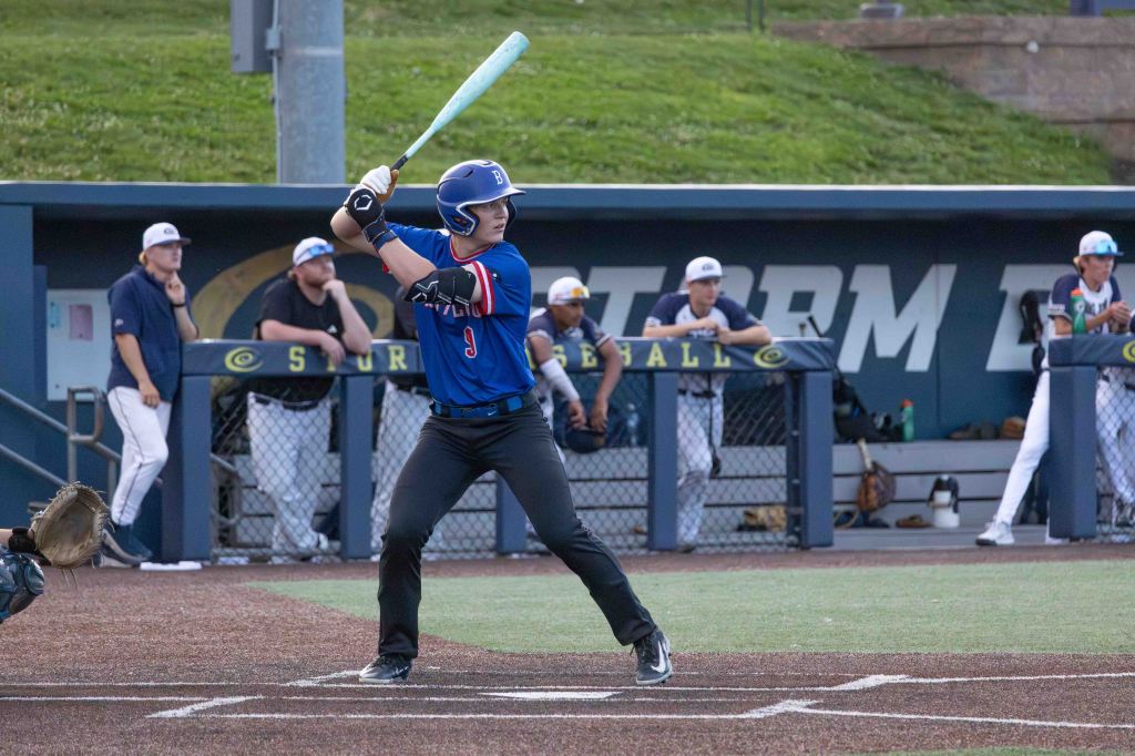 A baseball player in a blue uniform with the number 9 prepares to bat on a field, while teammates observe from the dugout in the background.