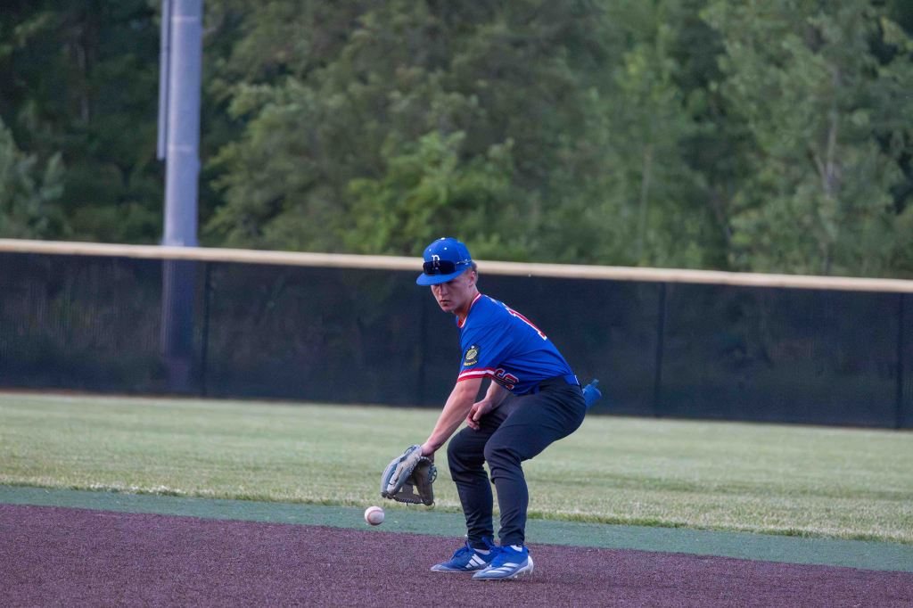 A young male baseball player in a blue jersey and cap is preparing to field a ground ball during a game, with a grassy outfield and trees in the background.