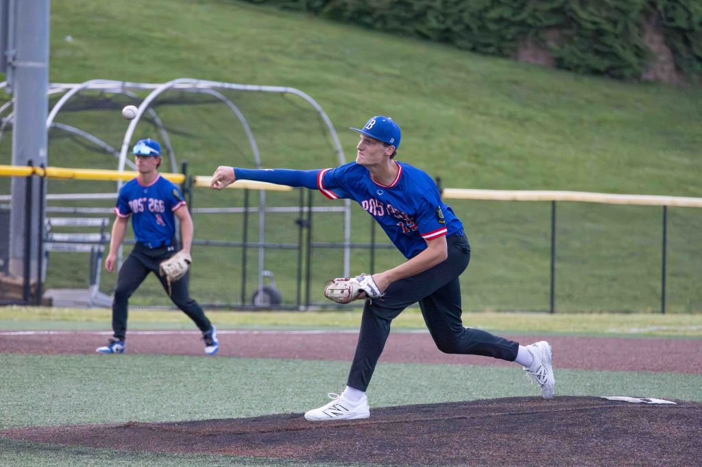 A young baseball pitcher in mid-throw, wearing a blue jersey and cap, with a batter in the background preparing to hit.