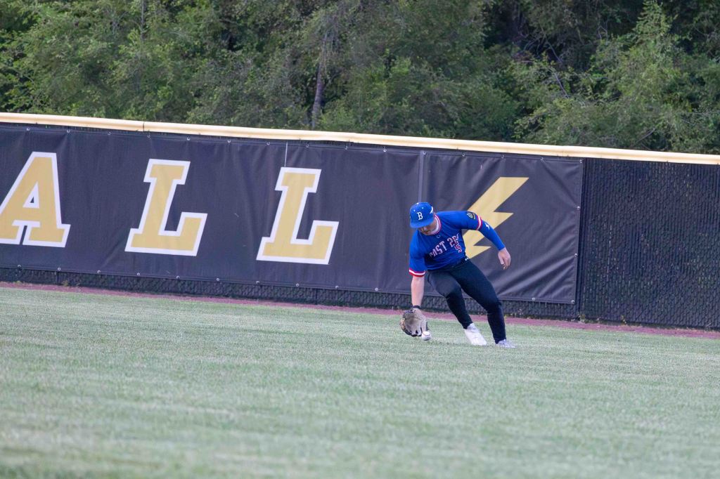 A baseball player in a blue jersey is crouching down to catch a ball on a grassy field with a black and yellow outfield wall in the background.