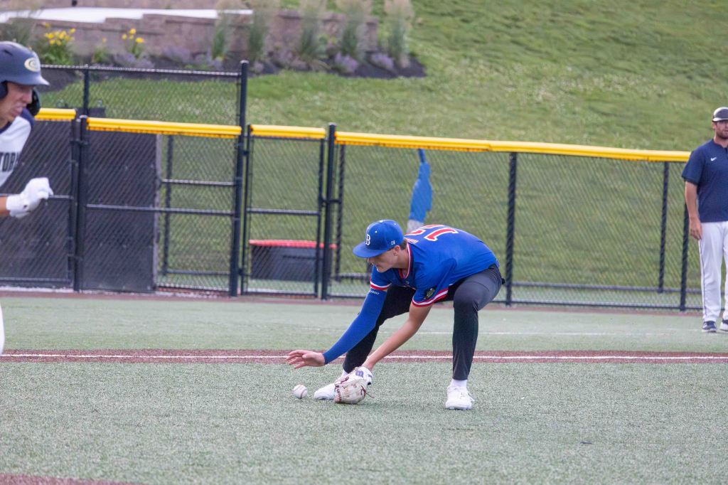 A baseball player in a blue jersey crouches to field a ground ball on a turf field, while a base runner in white approaches first base.