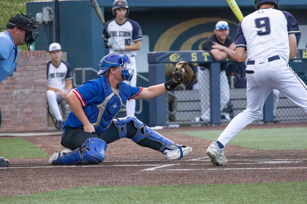 A baseball catcher in blue gear prepares to catch a pitch, with the umpire in blue behind him and players sitting in the dugout in the background.