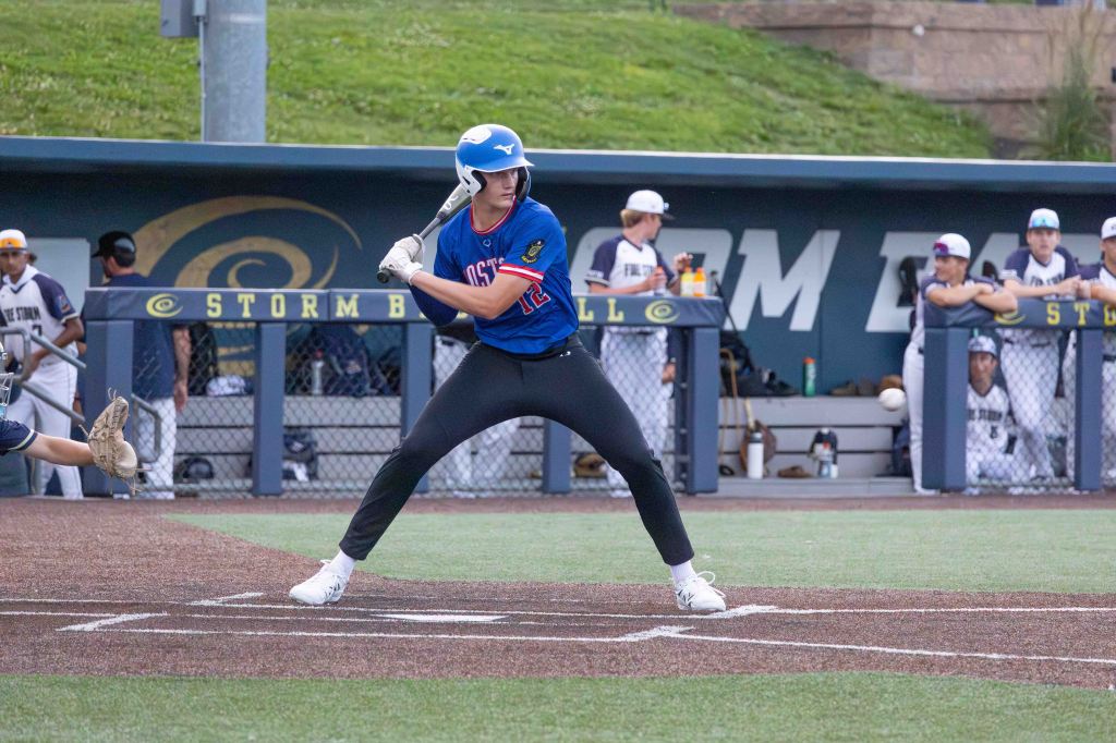 A baseball player in a blue jersey and white helmet stands in a batting stance on the field, preparing to hit a pitch. In the background, teammates watch from the dugout.