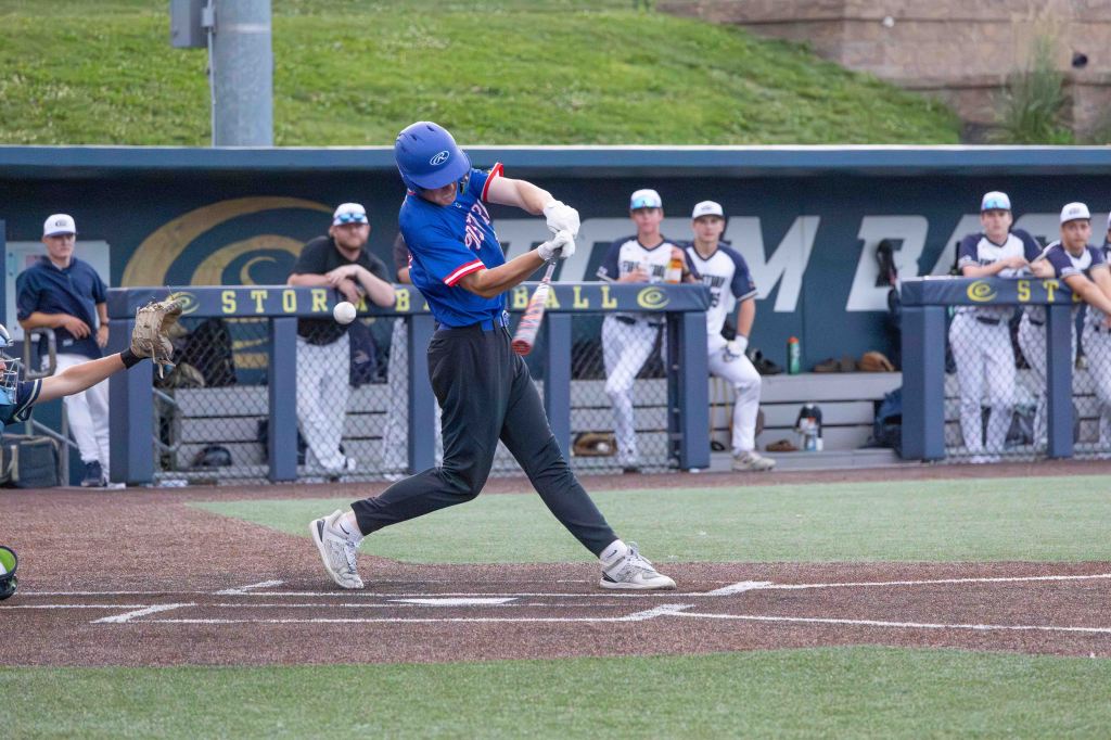 A young baseball player swings at a pitch during a game, while teammates observe from the dugout in the background.