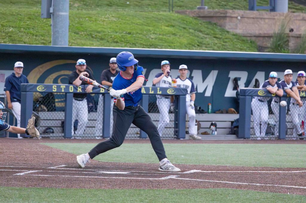 A baseball player swings a bat while attempting to hit a pitch during a game, with teammates and coaches visible in the dugout in the background.