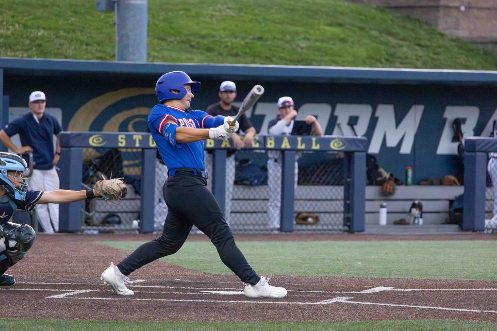 A baseball player in a blue jersey swings a bat at home plate, while a catcher in gear receives the pitch behind him. Teammates are visible in the dugout in the background.