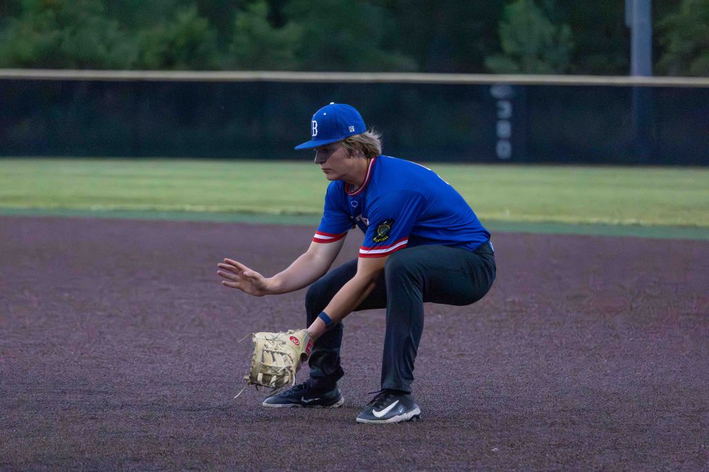 A young baseball player in a blue jersey and cap squatting on the field, preparing to field a ball on a dirt surface.