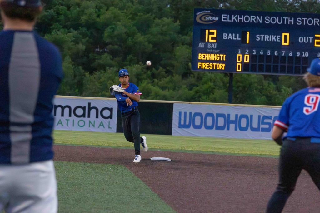 A player in a blue baseball uniform throws the ball at a game, with a scoreboard showing the current match statistics in the background.