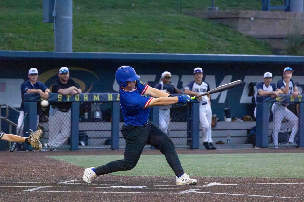 A baseball player in a blue jersey and cap swings a bat, making contact with a pitch, while teammates watch from the dugout.