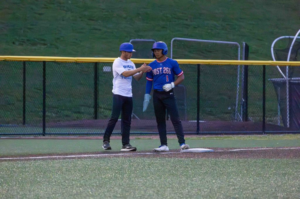 Two baseball players engaged in conversation on the field, one in a blue jersey and helmet, the other in a white shirt and black pants.