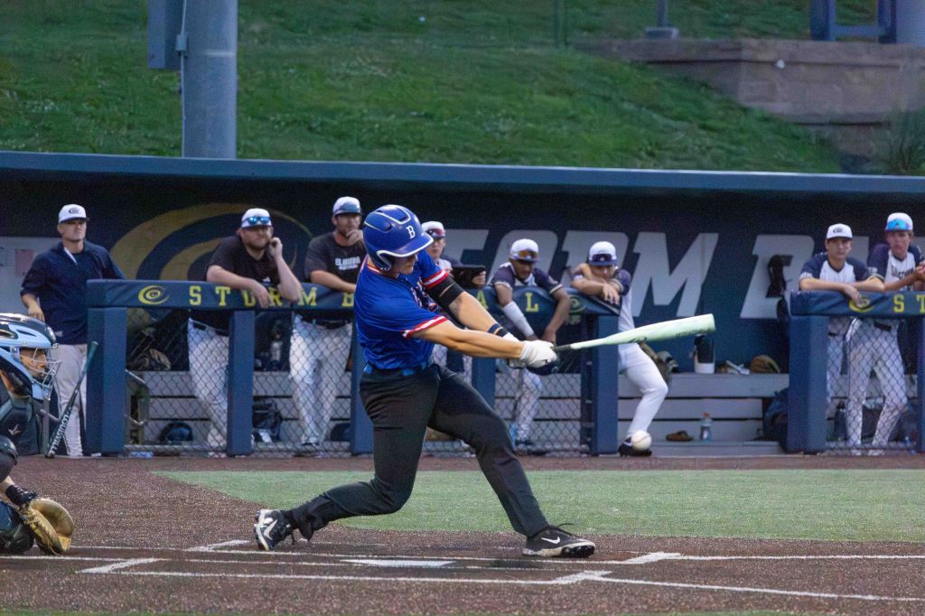 A baseball player swings a bat during a game, capturing the moment of contact with the ball, while teammates observe from the dugout.