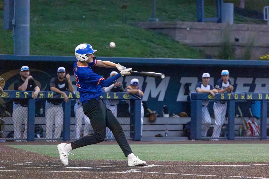 A baseball player in a blue jersey swings at a pitch while players watch from a dugout in the background.