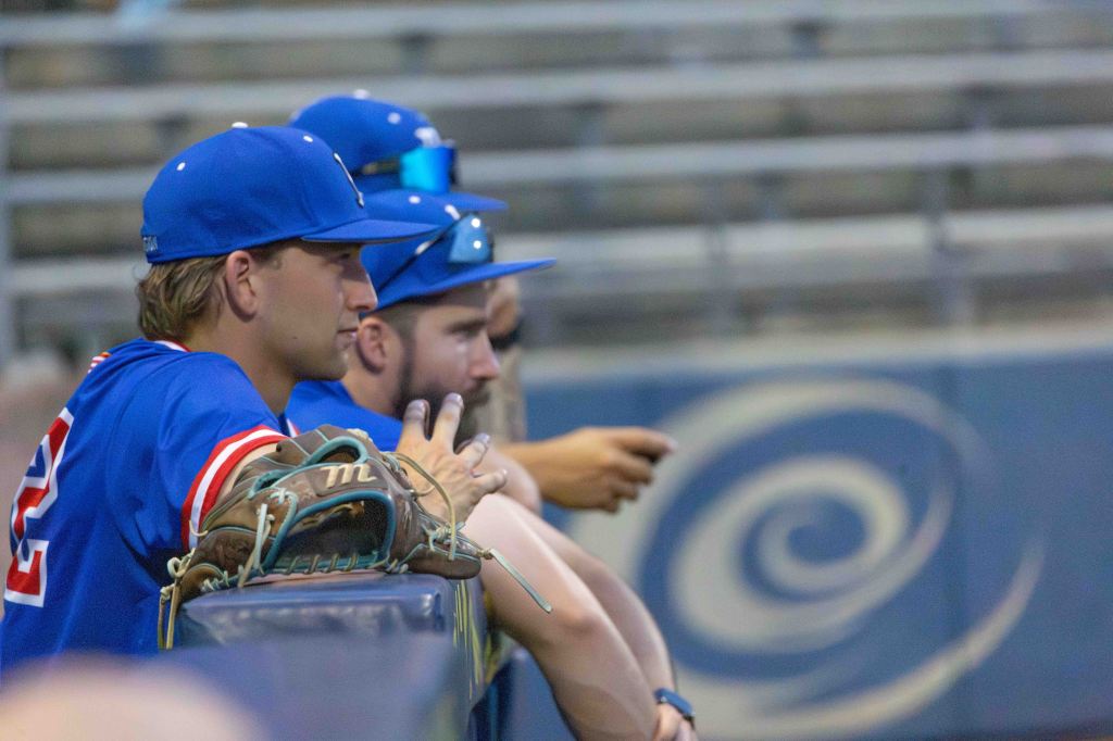 Three male baseball players watching from the dugout, wearing blue team uniforms and caps, with a baseball glove resting on the bench.