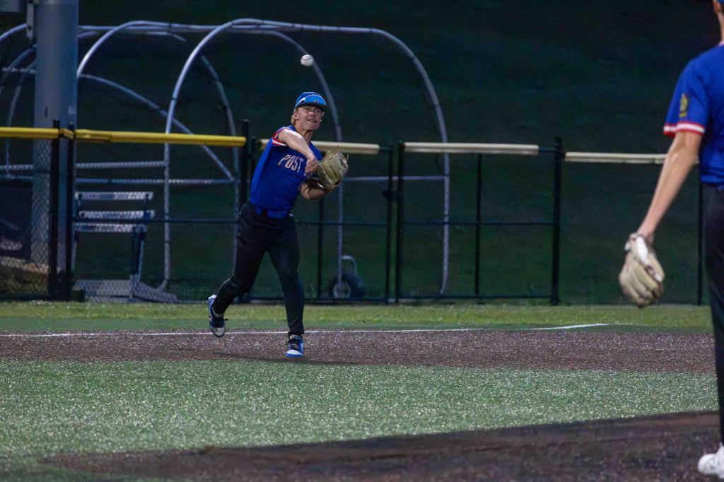 A baseball player wearing a blue jersey throws a ball towards a teammate during a game at night.