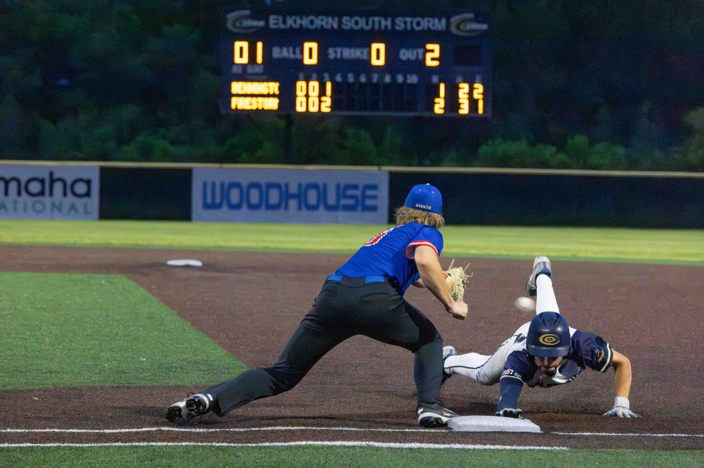 A baseball player in a blue jersey and cap reaches for the base while another player in black slides towards it, with a scoreboard showing the game status in the background.