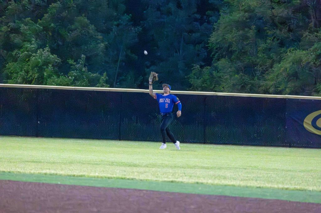 A baseball player in a blue jersey catches a fly ball in the outfield under evening lights.