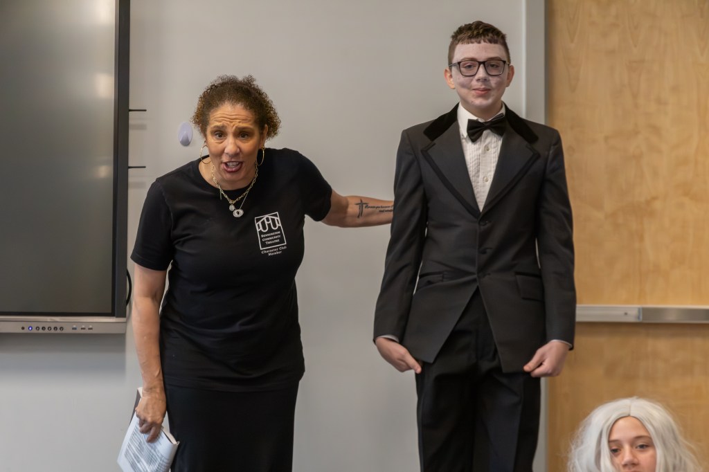 An engaged speaker in a black t-shirt is addressing an audience while standing next to a young man in a tuxedo with glasses. A person with white hair is visible in the foreground.
