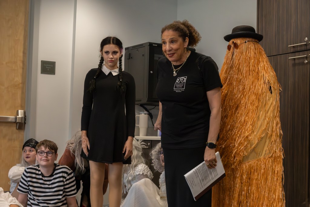 A group of children dressed in costumes stand together, with one girl in a black dress and braids looking serious. An adult woman in a black t-shirt talks to the group, and a character in a straw-like costume stands beside her.