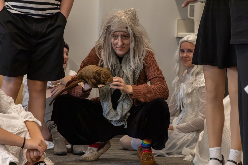 A person in a creative costume with long gray hair squats and holds a stuffed toy, surrounded by others in various outfits, in a friendly gathering.