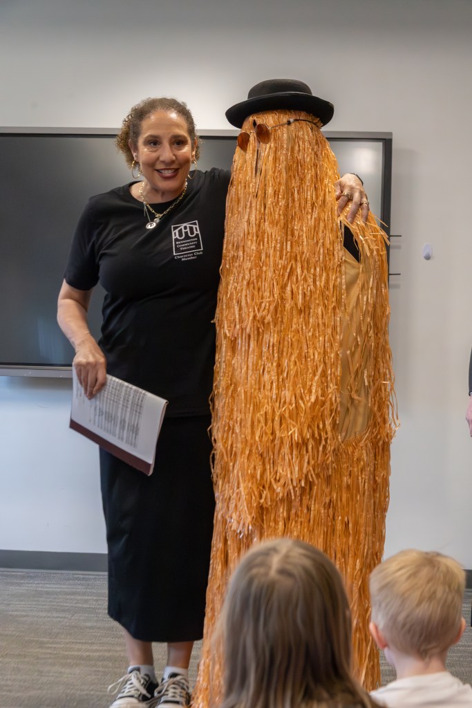 A woman stands next to a tall, costumed figure resembling a shaggy orange creature, both smiling. The woman holds a paper and wears a black shirt. The backdrop features a digital screen.