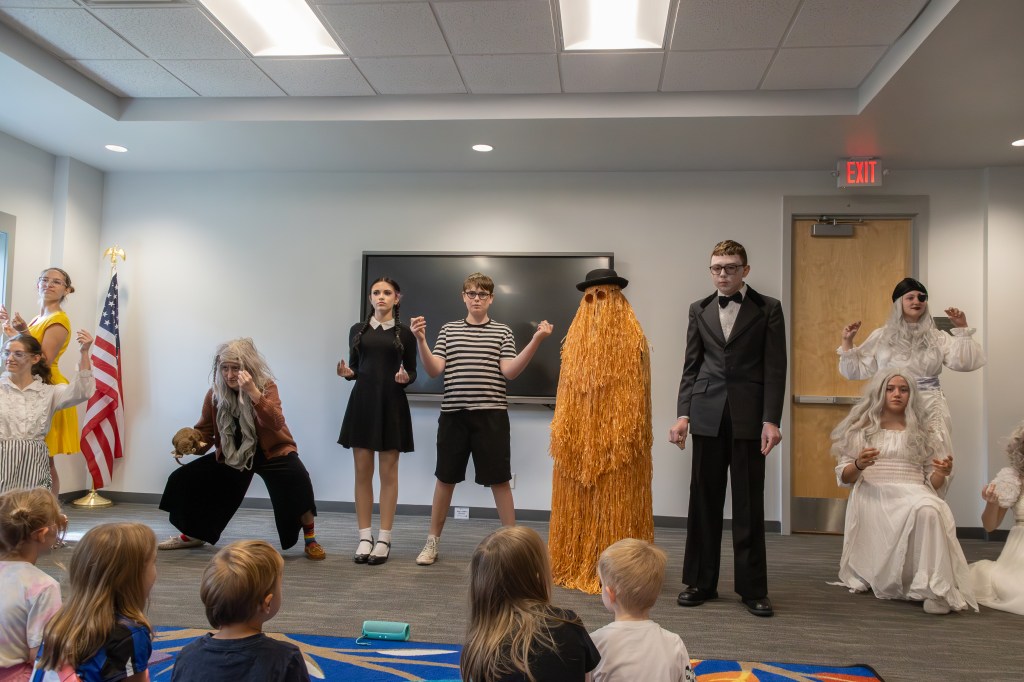 A group of children performing a play in costumes, featuring characters from a story. The scene includes various characters like a witch, a ghost, and a creature with an orange shaggy costume. The audience, consisting of young children, is seated in front, watching the performance.