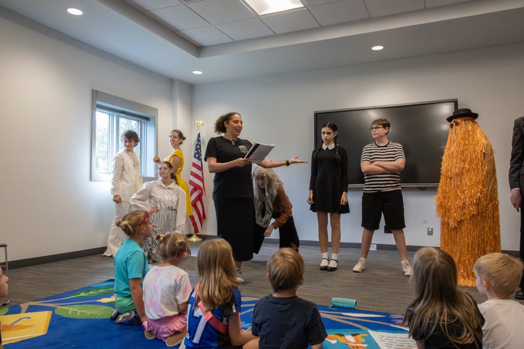 A group of children and adults performing a theatrical scene in a classroom, with one adult reading from a script, while various characters, including a person in a fringed costume, stand in front of an audience of children sitting on a colorful rug.