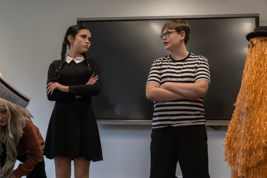 A dramatic scene featuring two young actors in a classroom setting. One actor is dressed in a black dress with braids, while the other wears a striped shirt and glasses. A third actor is crouching in the background, partially obscured.