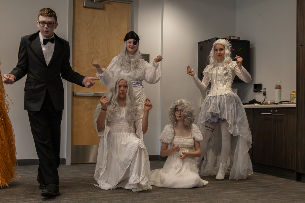 A group of five individuals dressed in elaborate costumes gathered in a room. The person on the left wears a tuxedo, while the others wear white dresses, some with long hair or wigs, striking poses.