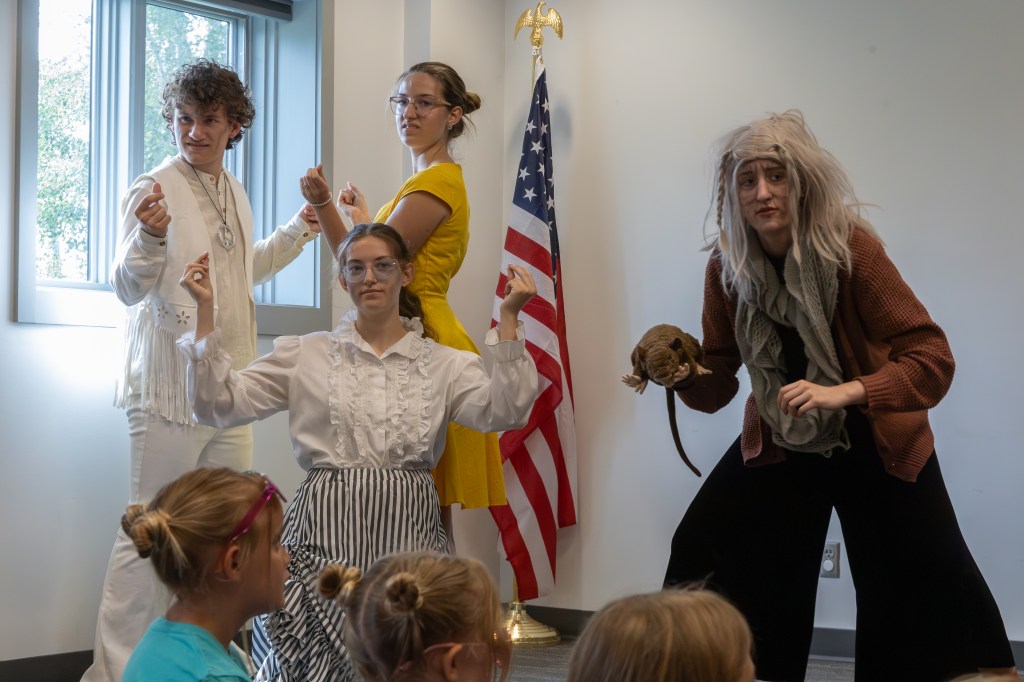 A group of four performers in costumes posing in front of an American flag, with two holding props. One performer wears a white outfit, while another features a vintage dress. A third performer appears in a sweater and holds a prop resembling a rat, with a character showing a playful expression.