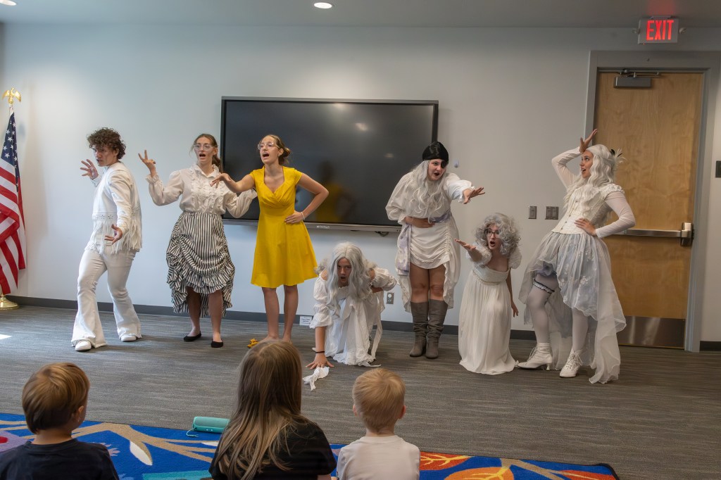 A group of six performers in theatrical costumes, including a girl in a yellow dress and several in white outfits, are acting on stage in front of an audience of children. The backdrop features an American flag and a large screen.