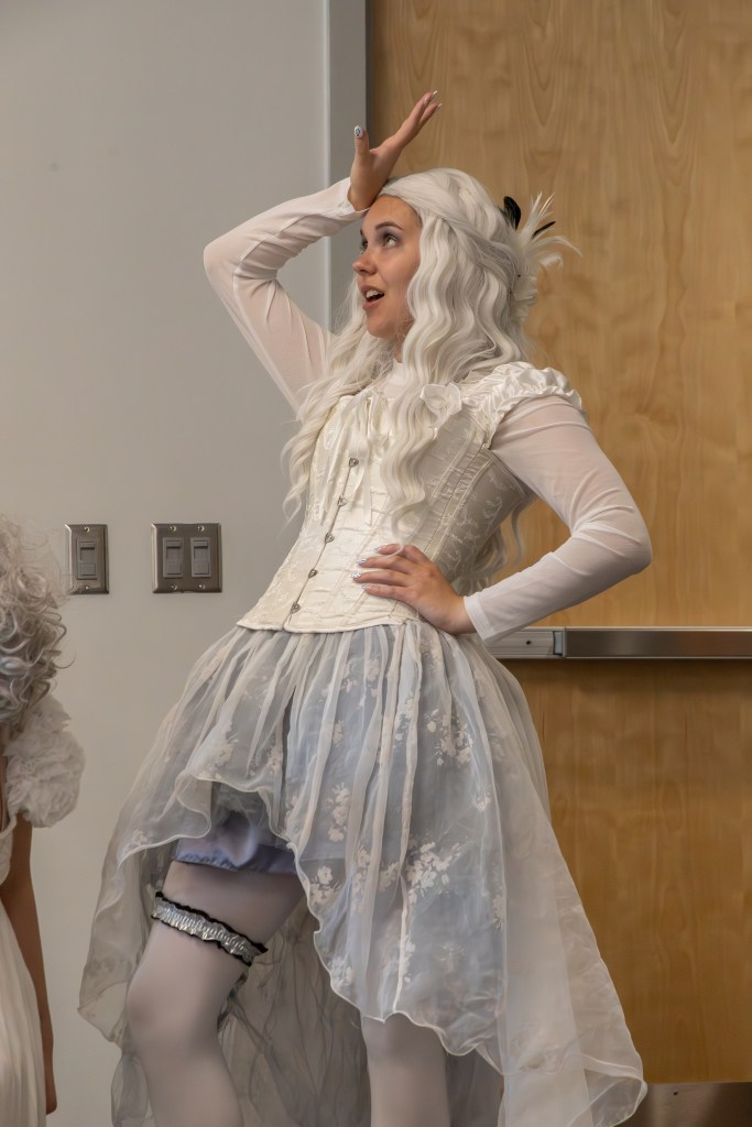 A person with long, wavy white hair wearing a white corset and flowy skirt strikes a playful pose, raising one hand to their forehead. The outfit includes a garter and is complemented by sheer white stockings.