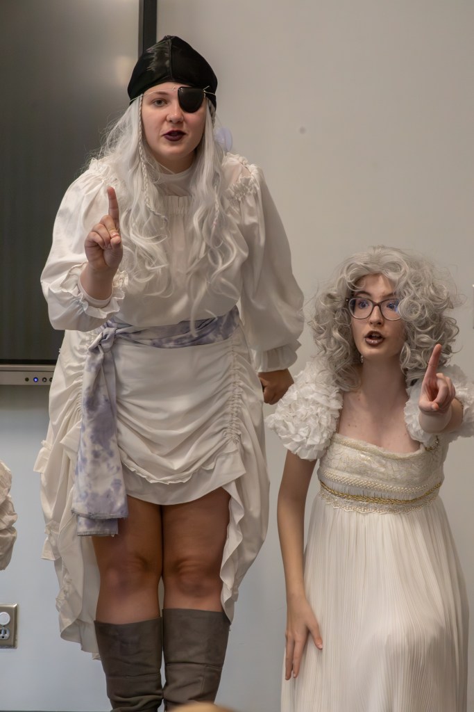 Two performers in costume, one wearing a pirate hat and eye patch, and the other with curly hair in a white dress, both striking dramatic poses.
