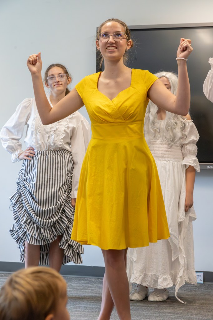 A girl in a yellow dress performs on stage with confidence, surrounded by other performers in historical costumes.