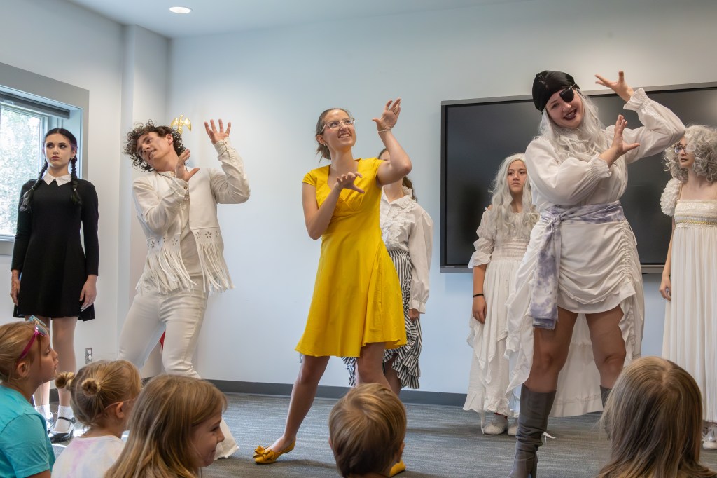 A performance scene featuring several actors in costumes, with a focus on a woman in a yellow dress leading the group. The actors display various poses, as a group of children watches them attentively.