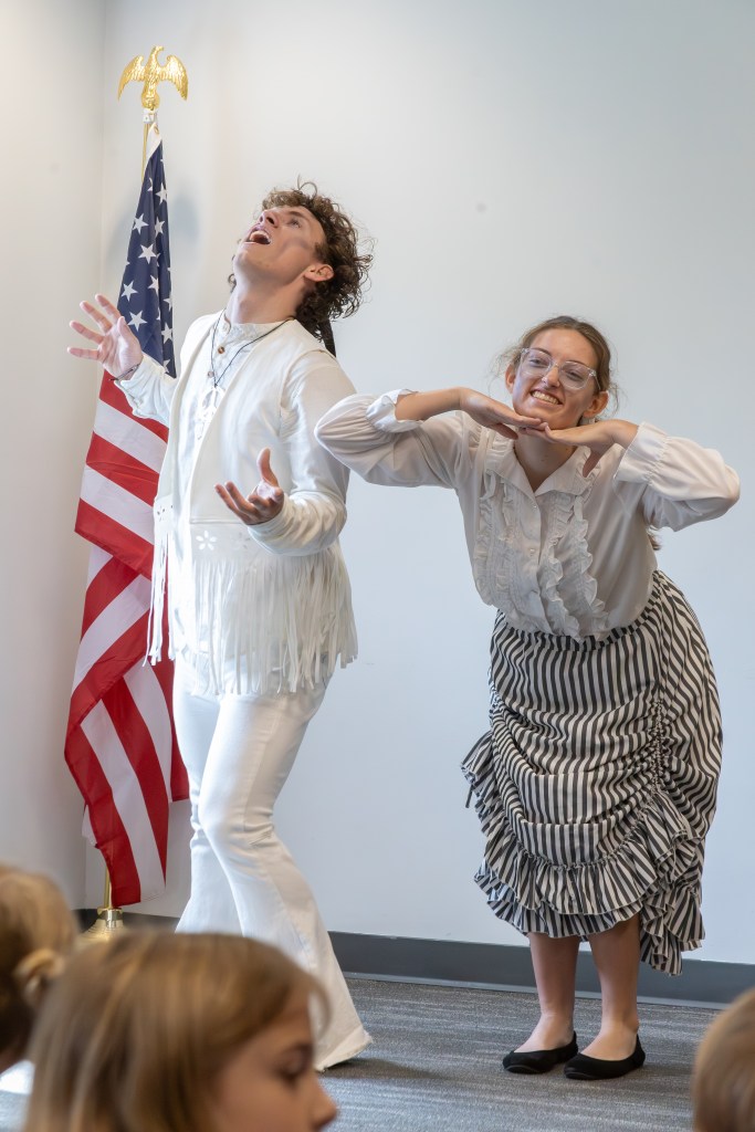 Two performers in a classroom setting striking playful poses, one in a fringed white outfit and the other in a striped skirt and white blouse, with an American flag in the background.