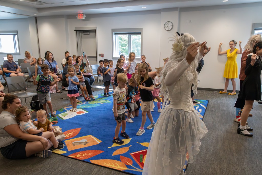 A lively children's event with kids and adults in an indoor space. Children are participating and interacting, some in costumes. A performer in a white dress engages with the audience on a colorful carpet. Several adults watch from seated positions.